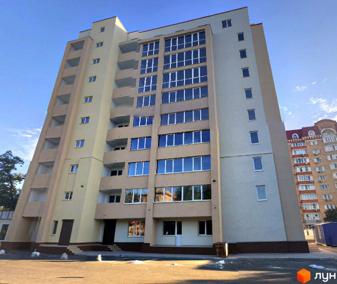 Багатоповерховий житловий будинок світлого кольору з великими вікнами та балконами. Прибудинкова територія з мощенням.