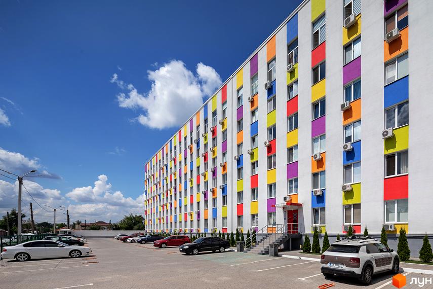 Житловий комплекс Бестужевські сади з яскравим фасадом у кольорову смужку. На прибудинковій території парковка з автомобілями та зелені насадження.