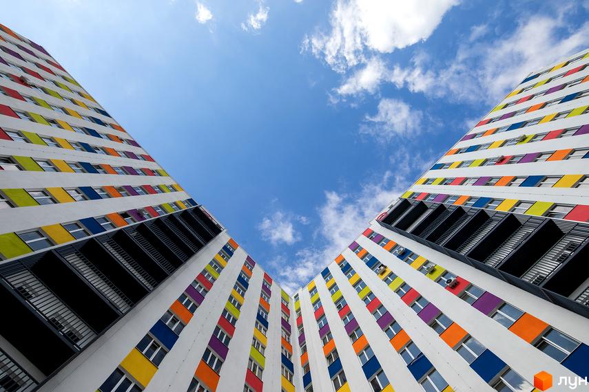 Зображення фасаду Новобудови Воробйовi гори з кольоровими вставками та балконами. Вид знизу вгору на тлі блакитного неба з хмарами.