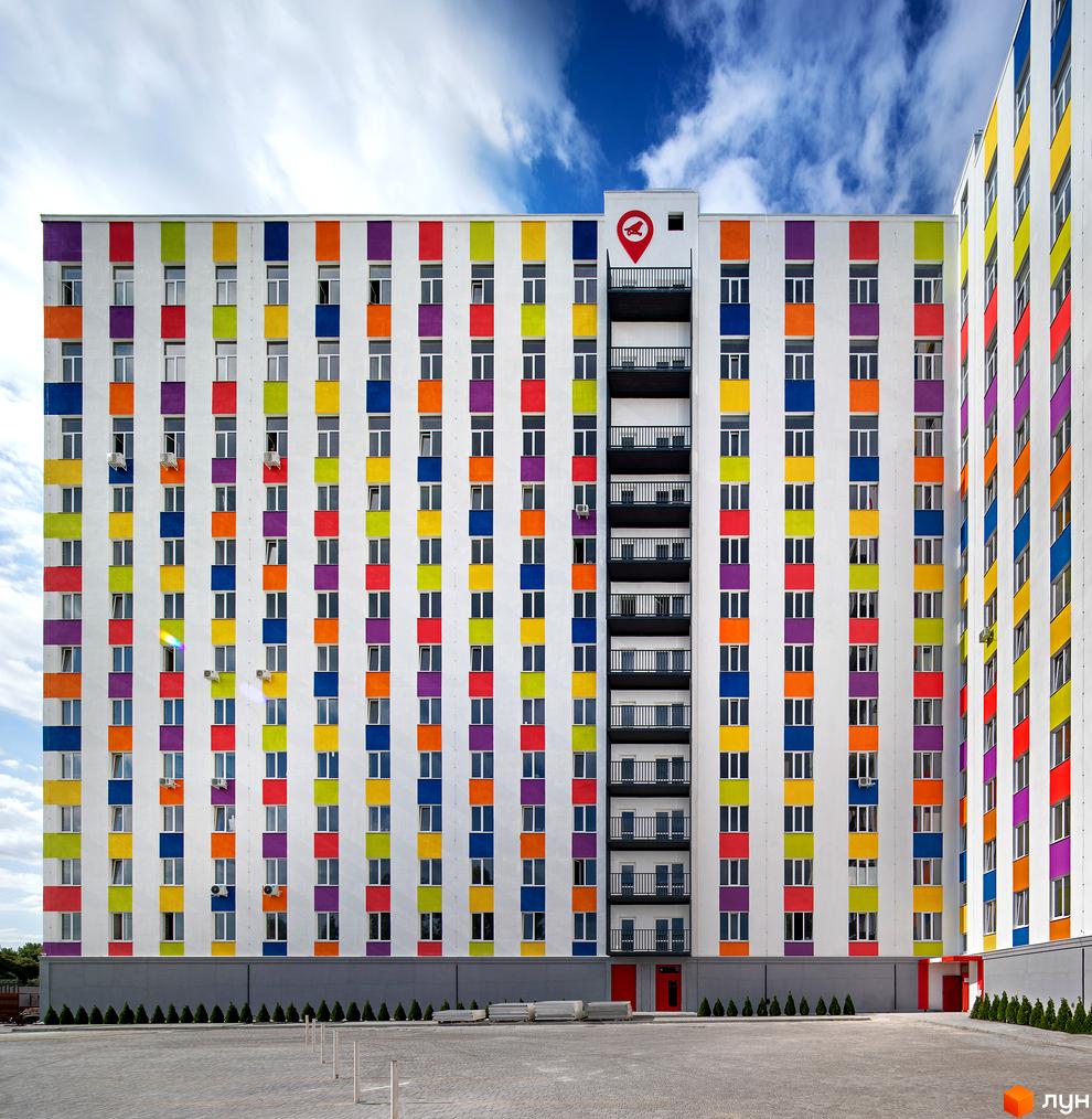 Яскравий багатоповерховий будинок з різнокольоровими смугами на фасаді. Поруч з будинком розташована прибудинкова територія з деревами та парковкою.