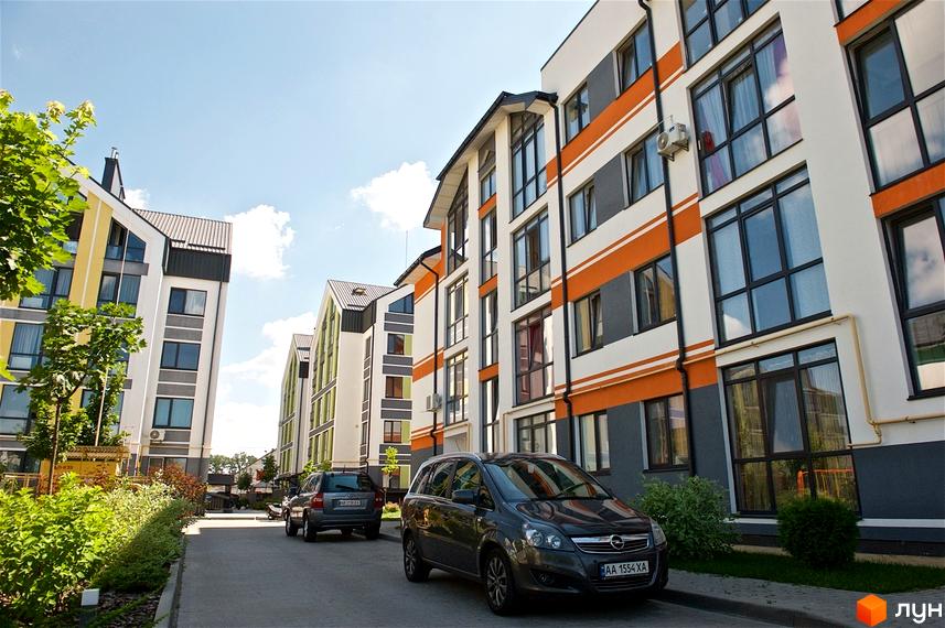 Новобудова ЖК Петрівське містечко з яскравим фасадом та панорамними вікнами. Прибудинкова територія з доглянутими зеленими насадженнями та автомобілями.