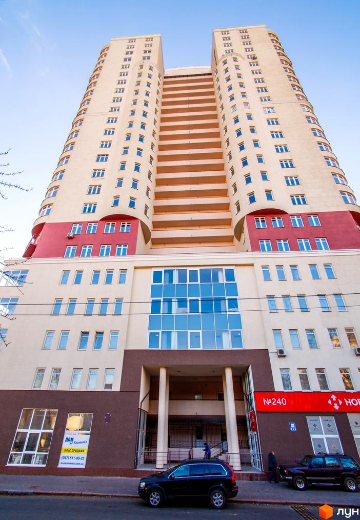 Житловий комплекс Будинок на Клименка: висока багатоповерхова будівля світлого кольору з червоними вставками та великими вікнами. На прибудинковій території видно автомобілі та пішоходів.