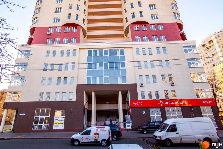 Житловий комплекс Будинок на Клименка: багатоповерховий будинок з бежево-червоним фасадом, великими вікнами та комерційними приміщеннями. На першому поверсі Нова Пошта та офіс продажу, перед будинком вулиця з автомобілями.