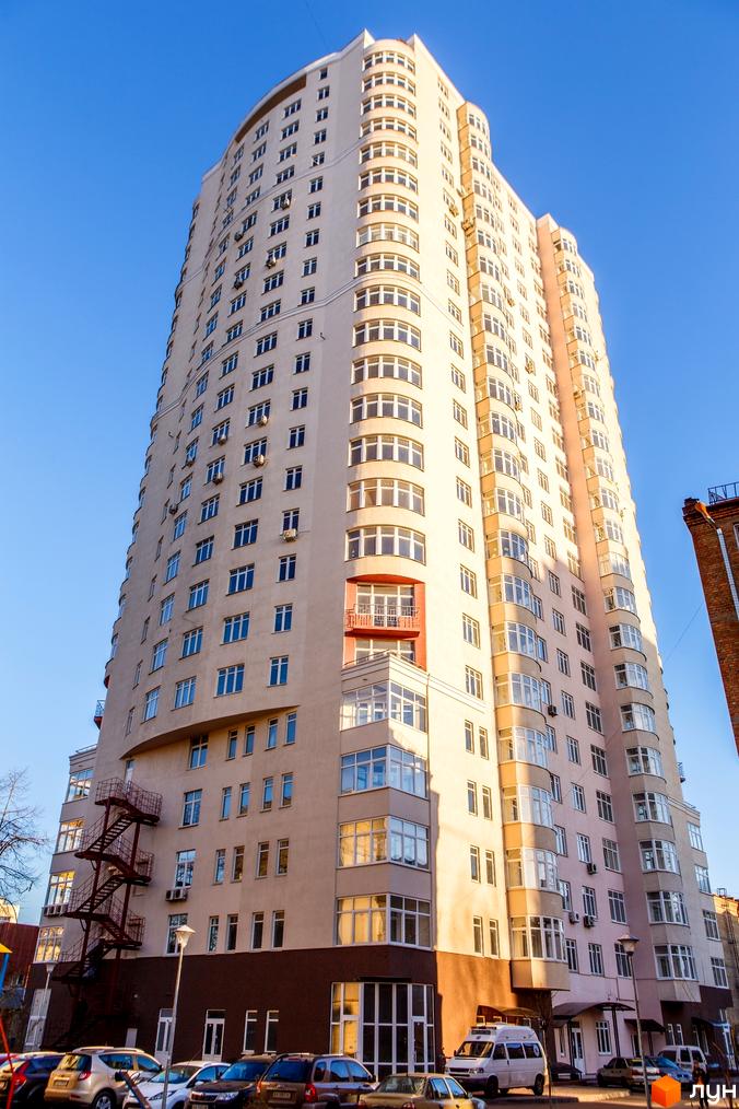 Житловий комплекс Будинок на Клименка: високий багатоповерховий будинок світлого кольору з численними вікнами та червоним балконом. Перед ним припарковані автомобілі.