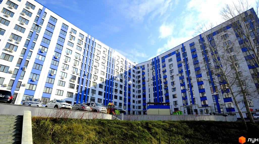 Житловий комплекс Рів'єра - багатоповерховий будинок з біло-синім фасадом та кондиціонерами на вікнах. На прибудинковій території розташовані автомобілі та дитячий майданчик.