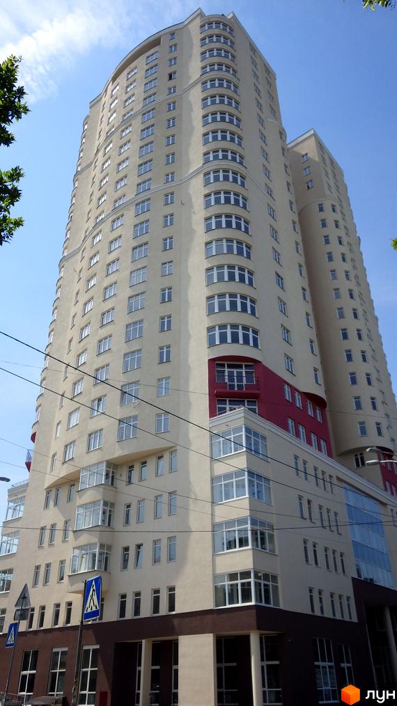 Високий житловий комплекс Будинок на Клименка кремового кольору з вигнутими фасадами та численними вікнами. На фасаді видно червоні вставки, а біля основи – пішохідний перехід.