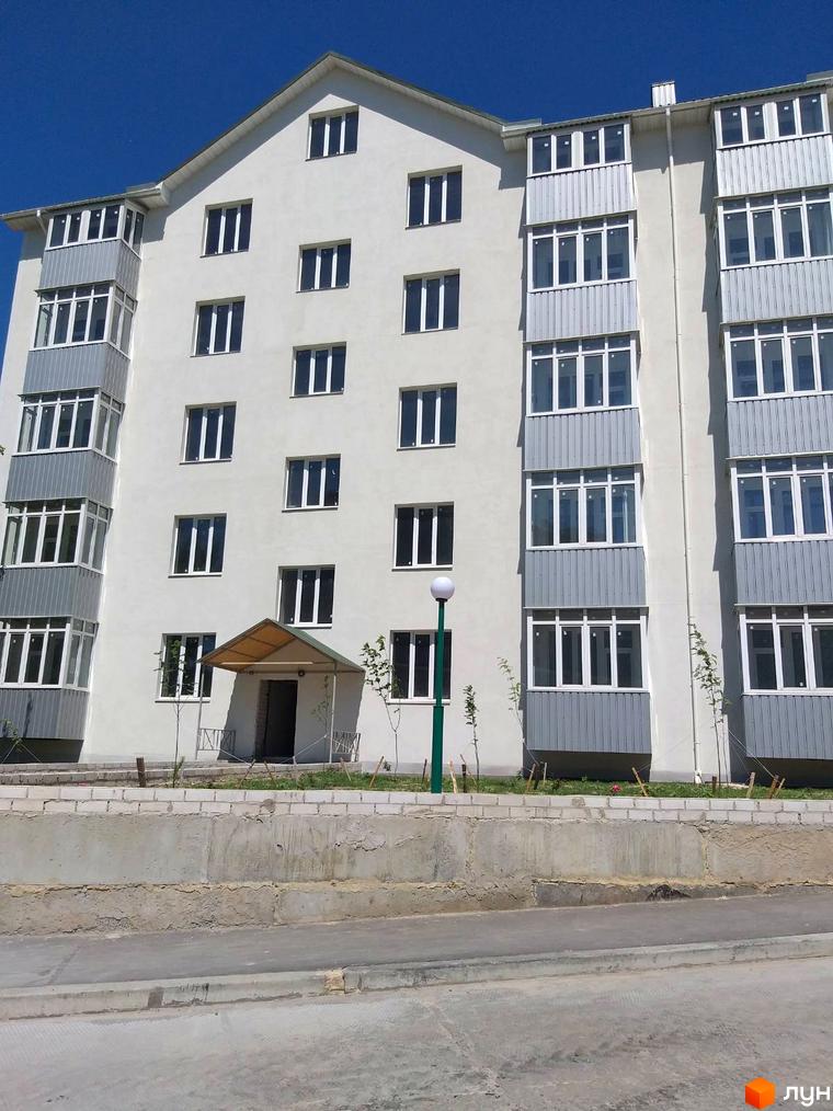 Фасад багатоповерхової новобудови світлого кольору з заскленими балконами та білими вікнами. Поруч бетонна огорожа і молоді дерева.
