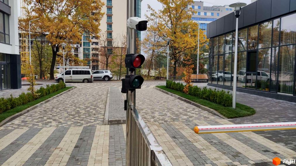 Житловий комплекс Містечко Липки: багатоповерхові будинки зі світлими фасадами. Прибудинкова територія з бруківкою, зеленню та шлагбаумом.