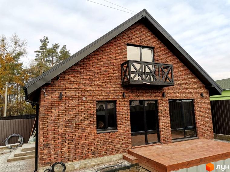 Цегляний будинок з двосхилим дахом, балконом та панорамними вікнами. На передньому плані дерев'яна тераса, навколо будинку бруківка та газон.