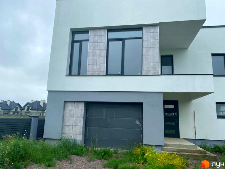 Фасад будинку Котеджного містечка OneSide House з білими та сірими стінами, великими вікнами, гаражем та вхідними сходами. Навколо будинку зелена трава та огорожа.