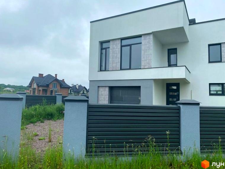 Фасад будинку Котеджного містечка OneSide House світлого кольору з темними вікнами та декоративними плитками. Перед ним – темний паркан та зелена трава.