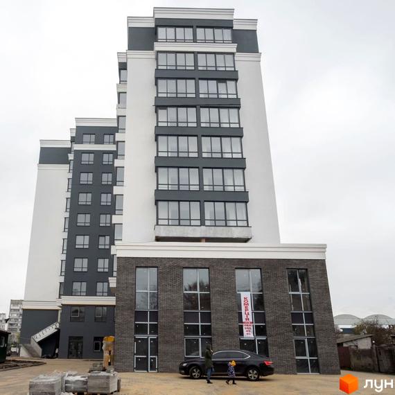 Фасад Житлового комплексу Олімп з білими та темно-сірими секціями, великими вікнами. На прибудинковій території видно автомобіль та будівельні матеріали.