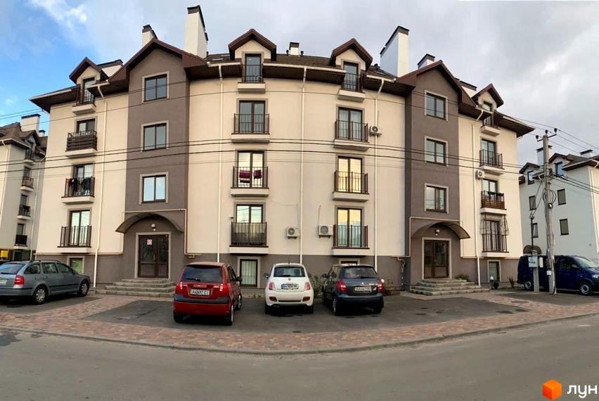 Зовнішній вигляд житлового комплексу Петрівський з автомобілями на парковці. Фасад будівлі світло-бежевий з сірими вставками, багато вікон та балконів.