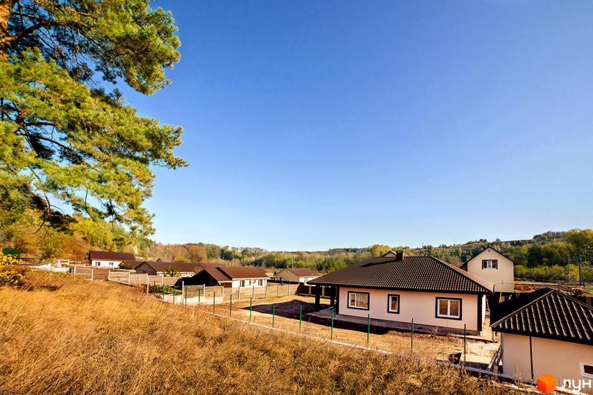 Котеджне містечко Лисичкина діброва. Зовнішній вигляд будинків з коричневими дахами та світлими стінами на тлі зеленого лісу та блакитного неба. Територія огороджена парканом, навколо суха трава.