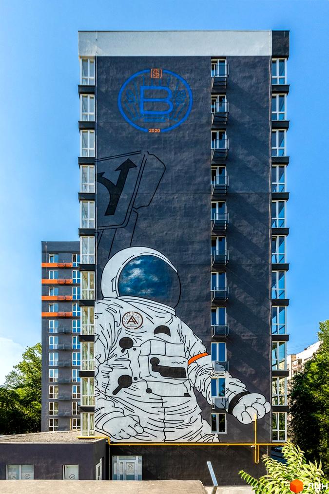 Зображення фасаду будинку Житловий комплекс Схід з муралом космонавта. Висотна будівля з великою кількістю вікон на фоні блакитного неба.