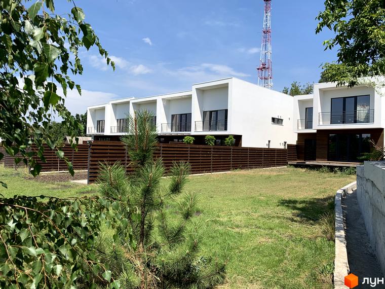 Таунхауси Green side: білі будинки з темними вікнами та балконами, деякі з дерев'яним оздобленням. Перед будинками зелений газон та дерев'яна огорожа.