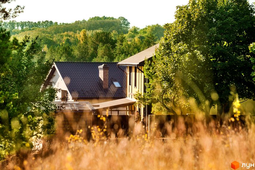 Котеджне містечко Лисичкина діброва. Будинок з коричневим дахом та цегляним димарем, оточений зеленню. Прибудинкова територія з деревами та травою.