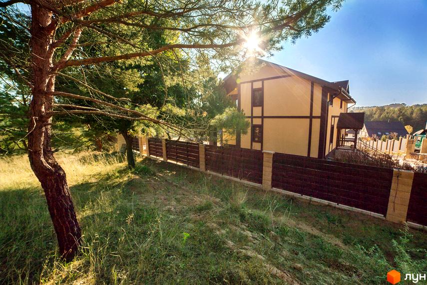 Котеджне містечко КМ Лисичкина діброва: двоповерховий будинок з коричневими балками та світлою стіною, огороджений парканом. Прибудинкова територія з деревами та травою.