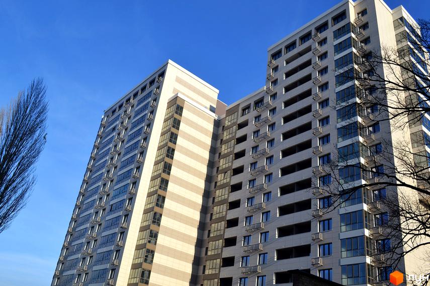 Вид на багатоповерховий житловий комплекс Островського, 40 з скляними фасадами та балконами на тлі блакитного неба. Будинок має сучасний дизайн з великою кількістю вікон.