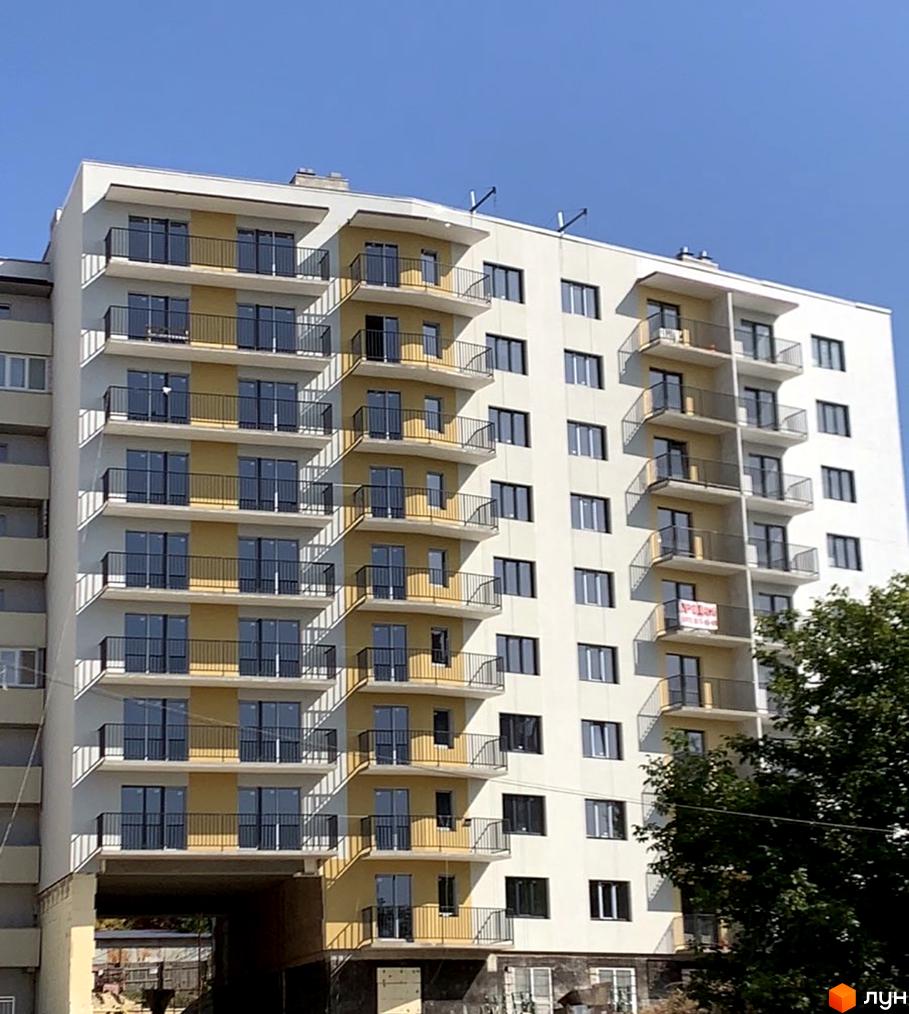 Новобудова на вул. Тарнавського, 7 - це багатоповерховий будинок з балконами та великими вікнами, що виходить на блакитне небо. Будинок має світлий фасад з жовтими вставками та чорними огорожами на балконах.