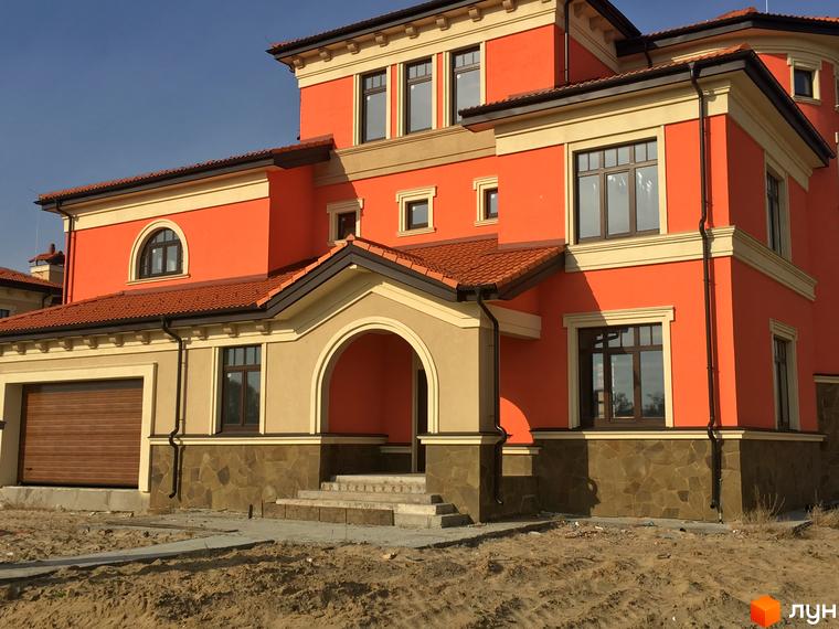 Котеджне містечко КМ Італійський квартал. Фасад будинку виконаний у помаранчевих та бежевих тонах з коричневим дахом та вікнами. Навколо будинку піщана територія.