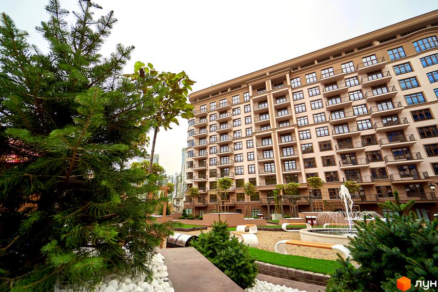 Висотна будівля житлового комплексу 52 Перлина (Pechersk Plaza) з балконами та терасами. На території комплексу розташовані зелені зони з деревами, лавками та фонтаном.