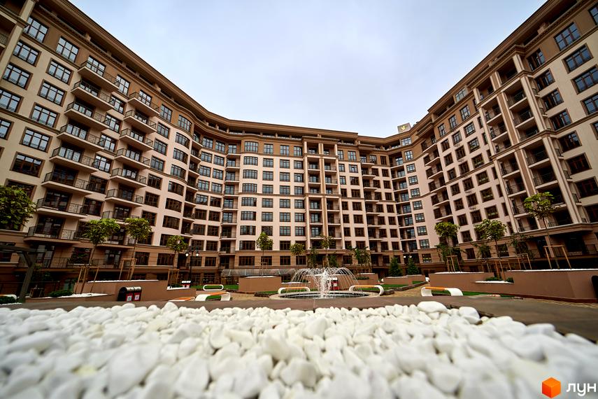 Житловий комплекс 52 Перлина (Pechersk Plaza). Висока будівля з балконами та великими вікнами. На передньому плані білі камінчики, позаду фонтан та лавки.