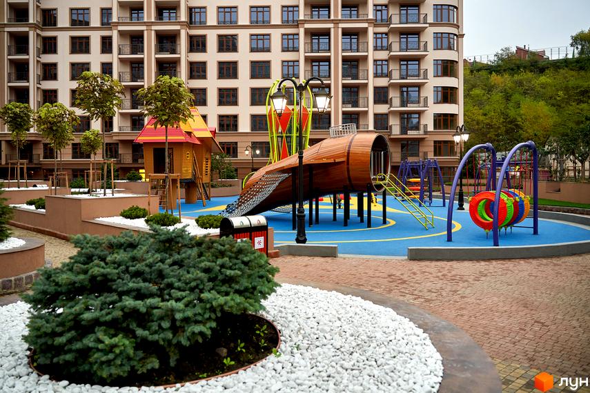 Будівля житлового комплексу 52 Перлина (Pechersk Plaza) з дитячим майданчиком на території. Фасад будинку з великою кількістю вікон, поруч дитячий майданчик з гіркою та гойдалками.