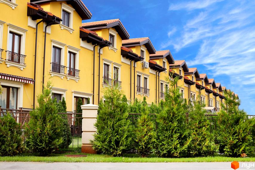 Зовнішній вигляд будинків КМ Італійський квартал, жовтий фасад, коричнева черепиця. Прибудинкова територія з зеленими туями та парканом.