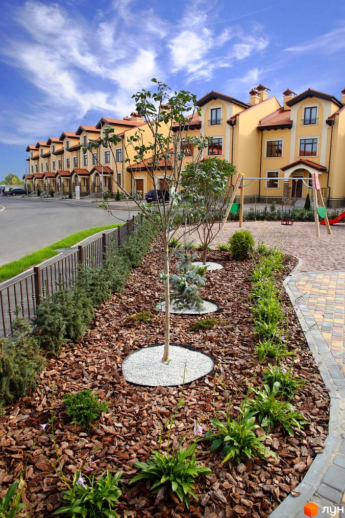 Котеджне містечко КМ Італійський квартал: жовті будинки з червоними дахами та доглянута прибудинкова територія з дитячим майданчиком. Зелені насадження та бруківка біля будинків.