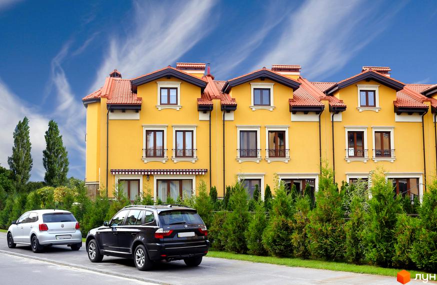 Котеджне містечко КМ Італійський квартал. Фасад жовтого кольору з червоним черепичним дахом. Перед будинком зелені туї та дві припарковані машини.