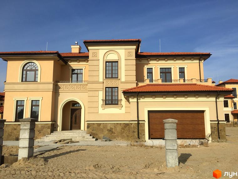 Котеджне містечко КМ Італійський квартал. Фасад будинку виконаний в бежевих тонах з червоною черепичною покрівлею. Навколо будинку піщана ділянка з елементами ландшафтного дизайну.