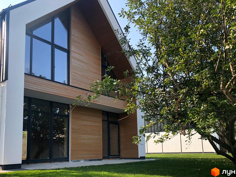 Котеджне містечко КМ Hill Park з дерев'яним фасадом та великими вікнами, зелений газон та дерево перед будинком.