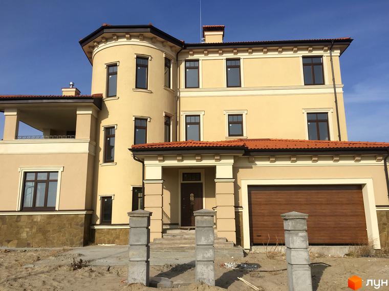 Котеджне містечко КМ Італійський квартал: двоповерховий будинок з жовтим фасадом, коричневим дахом та гаражними воротами. Навколо будинку піщана ділянка з бетонними стовпами.