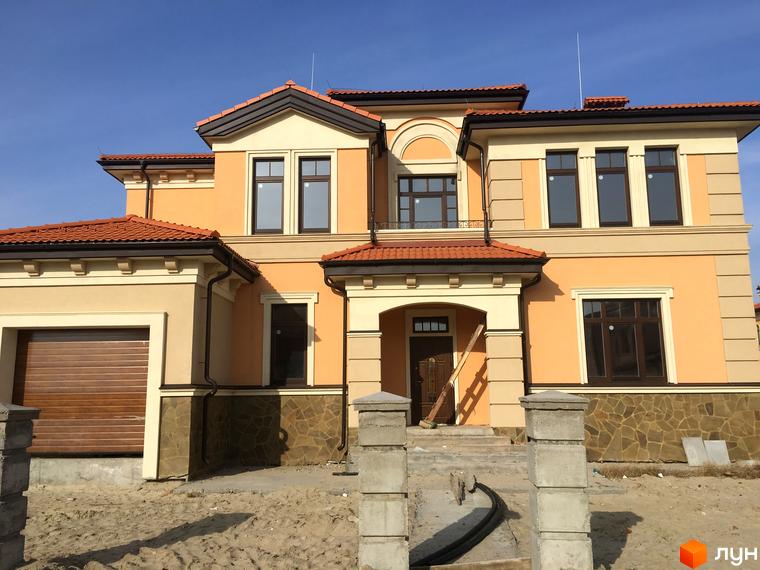 Котеджне містечко КМ Італійський квартал. Фасад двоповерхового будинку з червоною черепичною покрівлею, великими вікнами та гаражними воротами. Навколо будинку піщана ділянка з елементами ландшафтного дизайну.