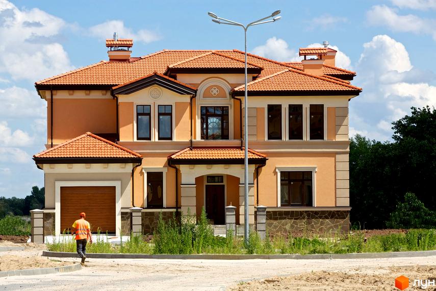 Котеджне містечко КМ Італійський квартал. Фасад двоповерхового будинку з помаранчевою черепичною кришею, великими вікнами та гаражними воротами. Навколо будинку зелені насадження та будівельні роботи.