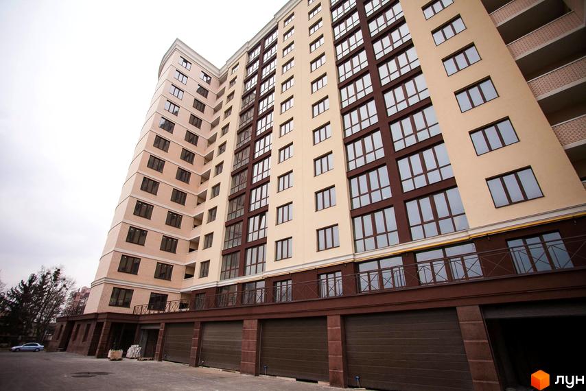 Багатоповерховий житловий комплекс Paradise з бежевими та коричневими фасадами, великими вікнами та балконами. На першому поверсі розташовані гаражі та асфальтований майданчик.