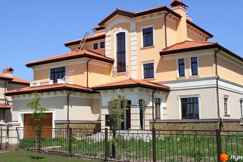 Котеджне містечко КМ Італійський квартал. Фасад будинку з помаранчевою штукатуркою, теракотовим дахом та великими вікнами. Огороджена територія з зеленою травою та деревами.