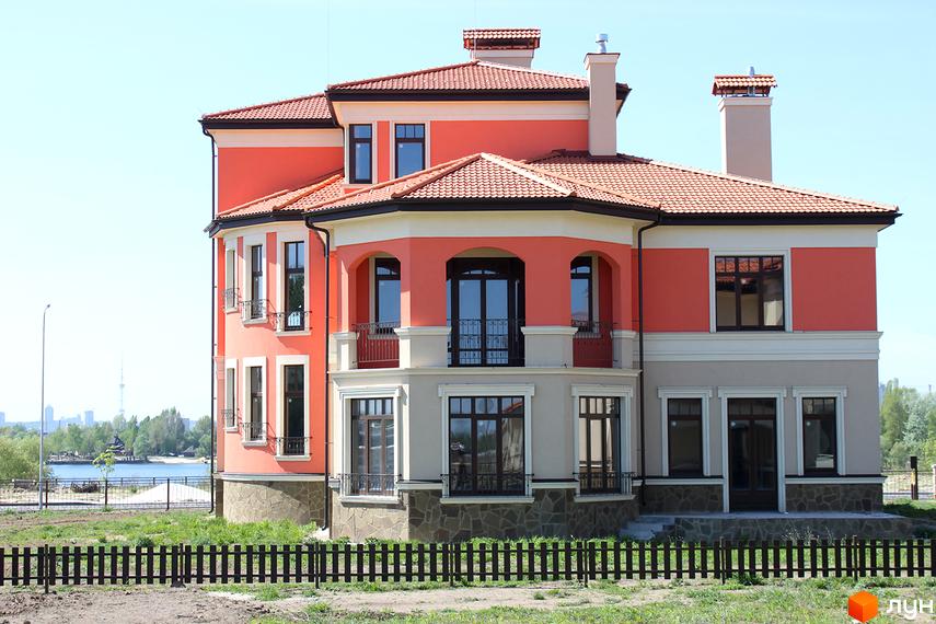 Котеджне містечко Італійський квартал: двоповерховий будинок з червоним дахом та фасадом персикового кольору, з балконами та великими вікнами. Навколо будинку зелена територія та чорний дерев'яний паркан.