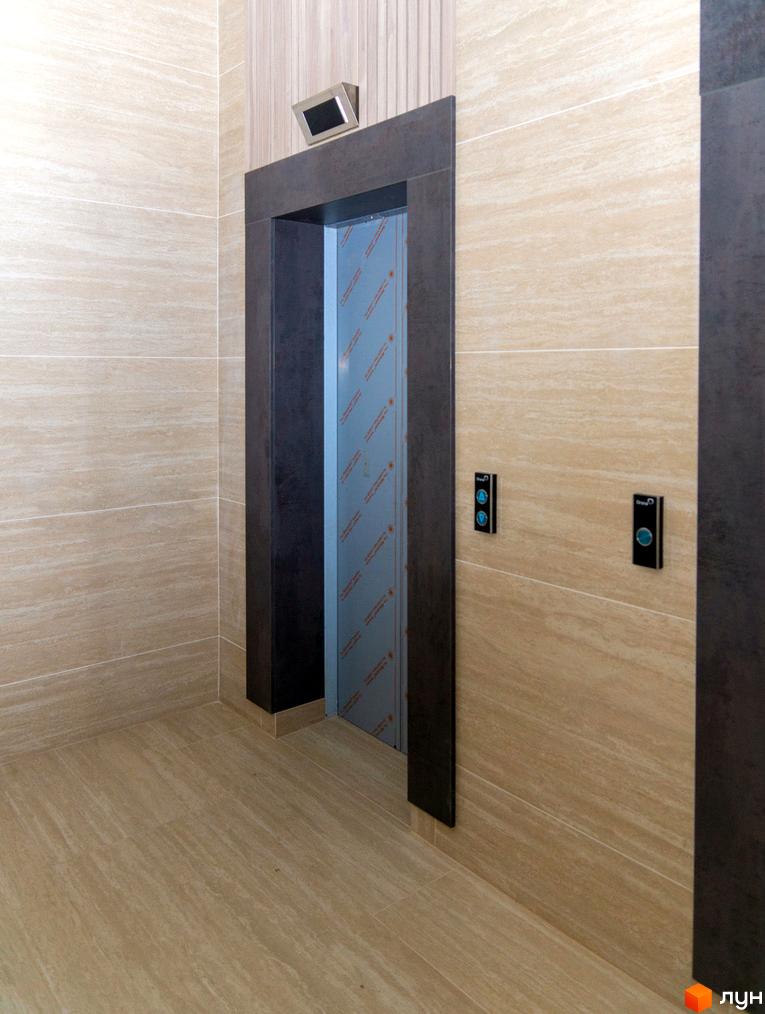 Інтер'єр холу з ліфтом у житловому комплексі Ярославів Град. Оздоблення стін та підлоги світлою плиткою під мармур, темний портал ліфта.