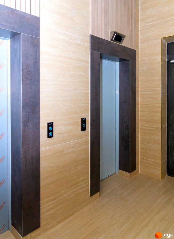 Два входи в ліфт з темними рамами та світлими стінами, оздоблені плиткою. На стінах біля входів розташовані кнопки виклику ліфта.