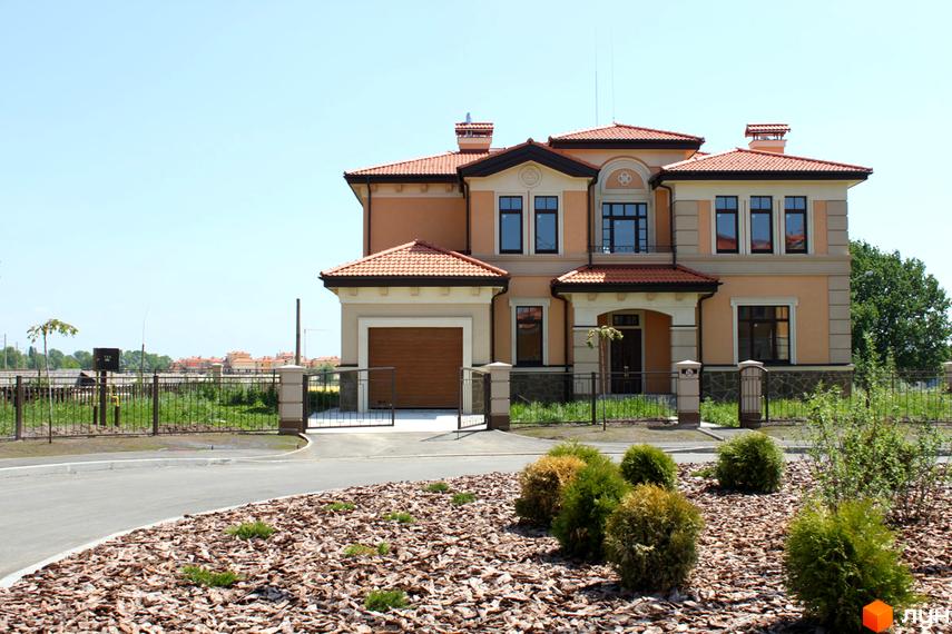 Котеджне містечко КМ Італійський квартал: двоповерховий будинок з червоною черепичною кришею та гаражем. Прибудинкова територія з доглянутими кущами та декоративною корою.