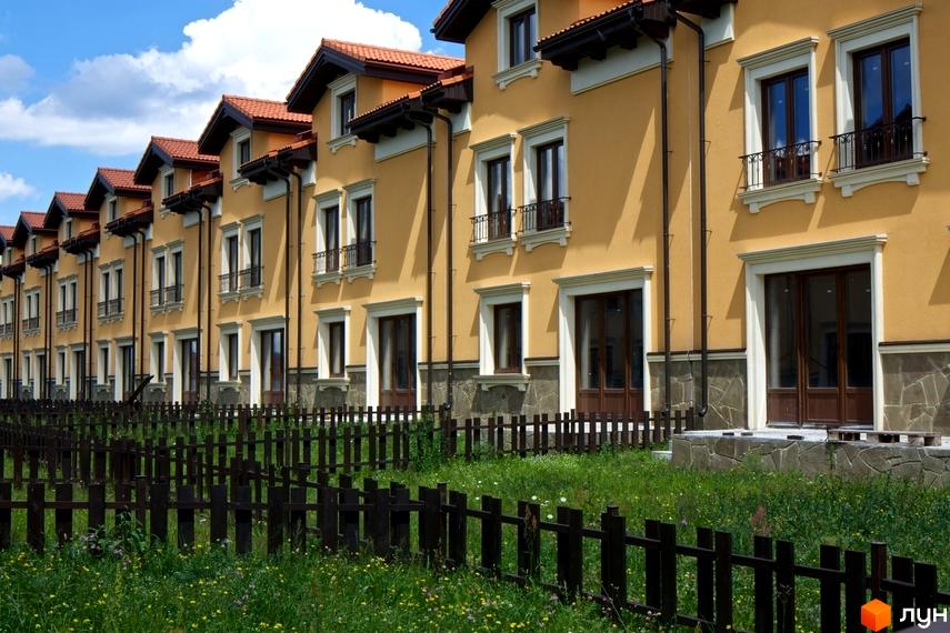 Зовнішній вигляд будинків КМ Італійський квартал з жовтим фасадом та червоним дахом. Попереду розташований дерев'яний паркан та зелена трава з квітами.