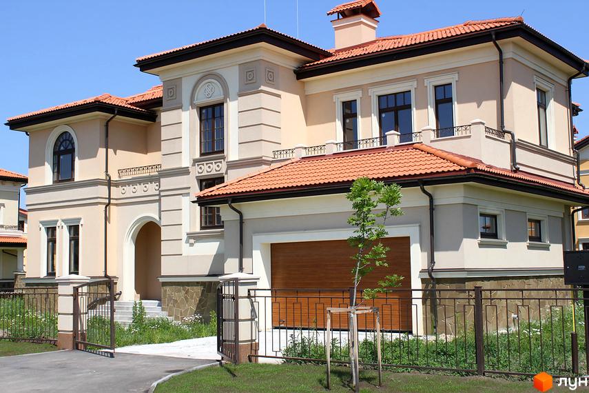 Котеджне містечко КМ Італійський квартал. Фасад двоповерхового будинку з червоним черепичним дахом, великими вікнами та гаражними воротами. Огороджена територія з доглянутим газоном та деревами.