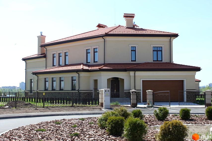 Котеджне містечко КМ Італійський квартал. Двоповерховий будинок світло-бежевого кольору з червоним черепичним дахом, великими вікнами та гаражними воротами. Навколо будинку доглянута прибудинкова територія з кущами та парканом.