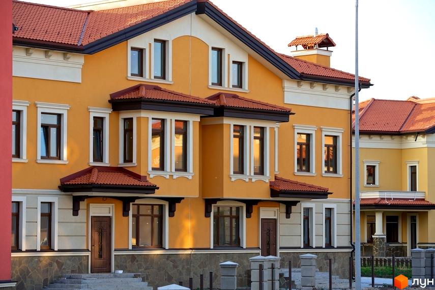 Котеджне містечко КМ Італійський квартал. Фасад будинку в жовтих тонах з червоною черепицею на даху. Навколо будинку облаштована прибудинкова територія з парканом та стовпчиками.