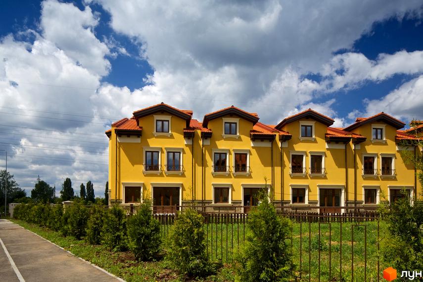 КМ Італійський квартал: фасади жовтих будинків з червоними дахами та вікнами. Прибудинкова територія з зеленими туями та парканом.