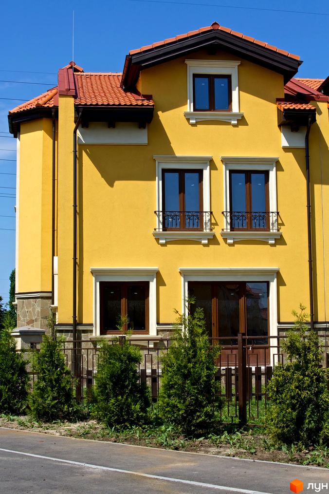 Житловий комплекс КМ Італійський квартал: жовтий двоповерховий будинок з коричневими вікнами та червоним дахом. Озеленена територія з туями та металевою огорожею.