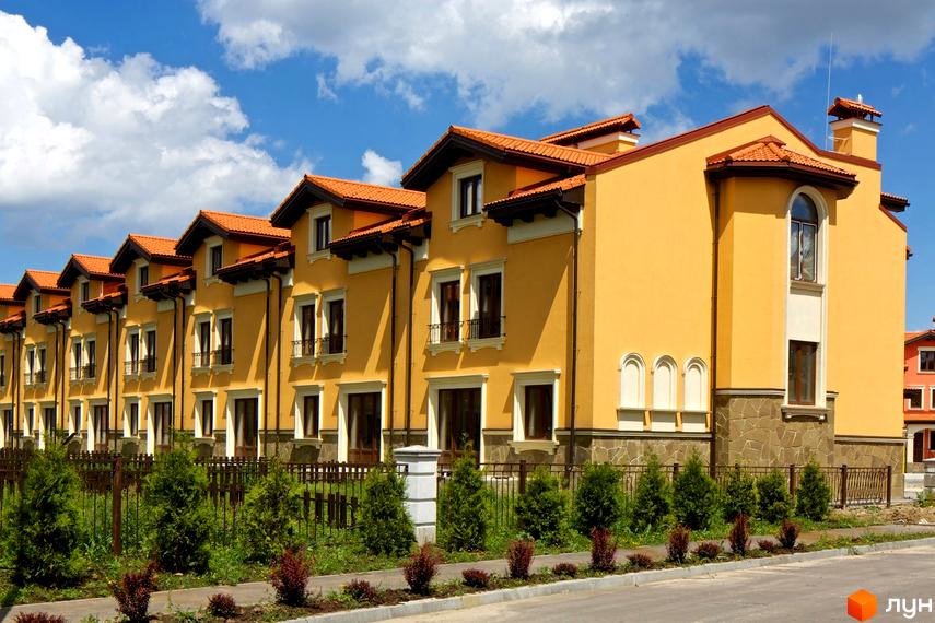 Котеджне містечко КМ Італійський квартал: жовті двоповерхові будинки з помаранчевими дахами, вікнами з кованими балконами та зеленими насадженнями. Затишна прибудинкова територія з огорожею та деревами.