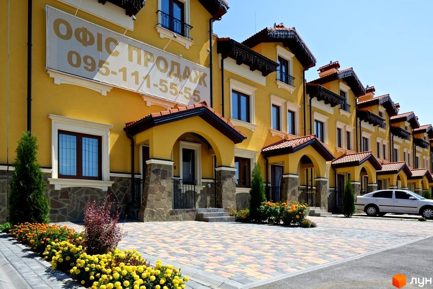 Зовнішній вигляд жовтих будинків КМ Італійський квартал з червоними дахами та кам'яним оздобленням цоколя. На прибудинковій території розташовані зелені насадження та квіткові клумби.